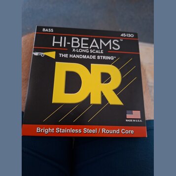 Fotografie znázorňuje čtvercové balení strun s nápisy BASS a 45-130. Na černém pozadí je výrazné žluté logo DR a texty HI-BEAMS™, X-LONG SCALE a THE…