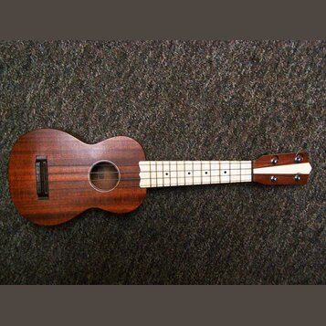 Hnědý dřevěný ukulele s hnedým korpusem a světlým hmatníkem. Uprostřed ozvučovacího otvoru je vidět štítek s nápisem "Kala". Nástroj vykazuje známky…