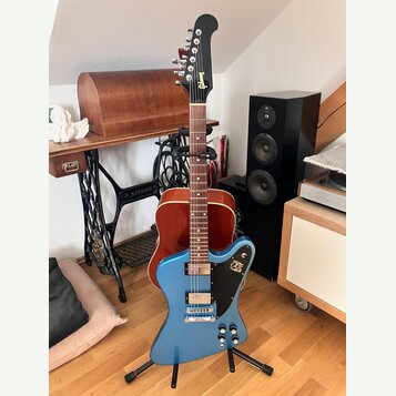 Modrá elektrická kytara s černým hmatníkem a hlavou, umístěná na černém stojanu. Na hlavě je bílý nápis Gibson. Tělo kytary má modrý lak, dva snímače…
