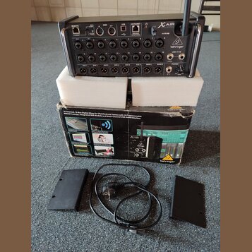 Digitální mixážní pult Behringer X AIR XR18 v černé barvě, umístěný na bílém polystyrenovém podkladu. Vedle přístroje je originální kartonová krabice…