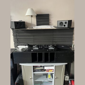 Na fotografii je set DJ vybavení umístěné na černých akustických panelech a bílém stolku. V horní části jsou dva černé studiové monitory, vprostřed č…