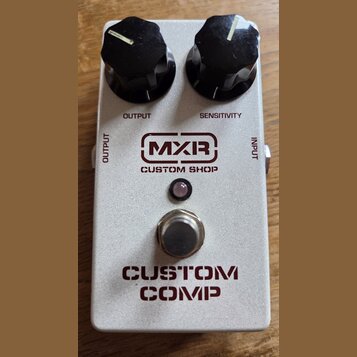 Stříbrný efekt pedál s nápisy MXR CUSTOM SHOP a CUSTOM COMP. Na horní straně jsou dva černé oválné ovládací knoflíky s popisky OUTPUT a SENSITIVITY.…