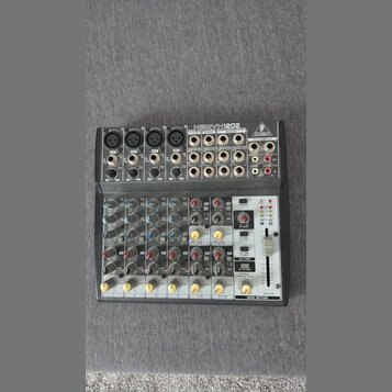 Šedý mixovací pult s nápisem XENYX 1202 a logem BEHRINGER. Zařízení obsahuje čtyři mikrofonní vstupy XLR, několik lineárních vstupů RCA a jack, poten…