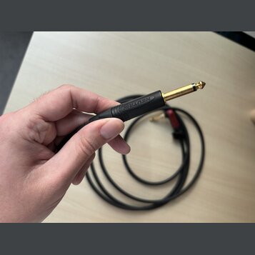 Ruka drží konec černého audio kabelu s pozlaceným 6,3mm jack konektorem. Na černém plastovém pouzdře konektoru je vyraženo logo NEUTRIK. V pozadí je…