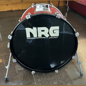 Černý basový buben s červeným korpusem, umístěný na kovovém stojanu. Na předním černém koženu je bílý nápis NRG DRUMS. Povrch nástroje vykazuje mírné…