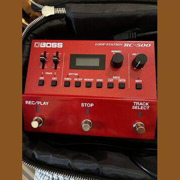 Červený looper Boss Loop Station RC-500 v černé brašně. Zařízení má digitální displej, několik ovládacích tlačítek a dva posuvníky. V dolní části jso…