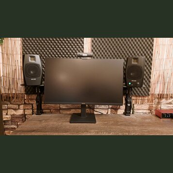 Na dřevěném stole stojí černý monitor značky SAMSUNG, po jeho stranách jsou umístěny dva černé studiové monitory ADAM AUDIO na černých stojánkách. Re…