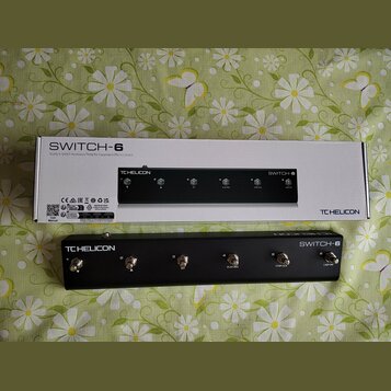 Černý kontrolní pedál s nápisy TC HELICON a SWITCH-6. Zařízení obsahuje šest stomečových spínačů s označením čísel 1 až 6 a doplňkovými nápisy HIT, P…