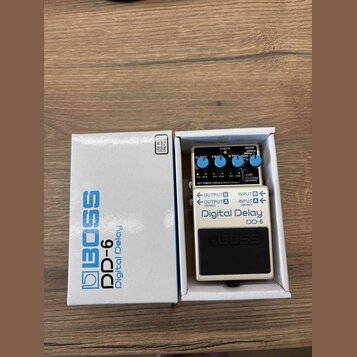 Na fotografii je efekt pedál Boss DD-6 Digital Delay v původním bílém kartonu. Pedál má stříbrné kovové tělo, černé ovládací knoflíky a nápisy Boss,…