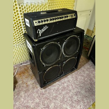 Černý kytarový zesilovač s hlavou umístěnou na basovém boxu. Na ovládacím panelu hlavy je nápis Fender a označení 1000. Box obsahuje čtyři reprodukto…