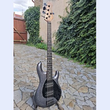 Elektrická basová kytara s tmavě šedým trpytivým povrchem těla a hnedým dřevěným hmatníkem a hlavou. Na hlavě je svisle napsaný nápis Ibanez. Tělo má…