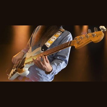 Bílá elektrická basová kytara s hnědým hmatníkem a světle dřevěnou hlavou. Na hlavě je vidět nápis Fender a text Jazz Bass. Nástroj má zlatý plech s…
