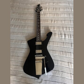 Černá elektrická kytara s atypickým, ostře hranatým tvarem těla a tmavým hmatníkem. Nástroj má dva humbuckery s kovovými kryty a stříbrný tremolo sys…