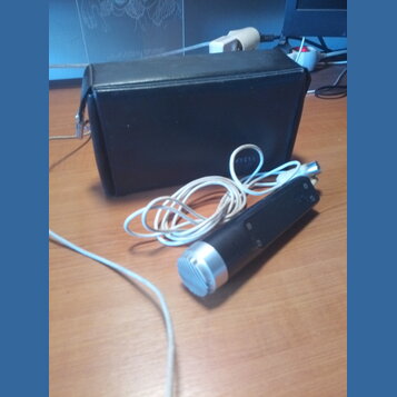 Na fotografii je černý mikrofon s kovovou stříbrnou mřížkou a bílým kabelem. Vedle mikrofonu je umístěno černé obdélníkové pouzdro z materiálu připom…