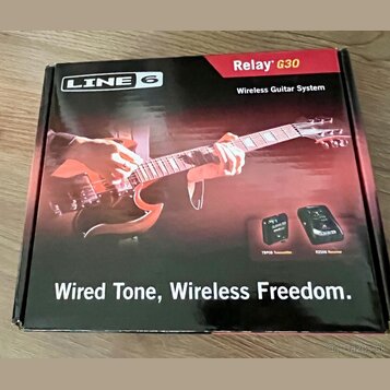 Kartonová krabice s nápisy LINE 6, Relay G30 a Wireless Guitar System. Na povrchu je fotografie rukou hrajících na červenou elektrickou kytaru a obrá…