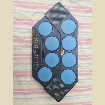 Elektronický bicí pad modré barvy s černým plastovým rámem. Na přístroji je nápis YAMAHA a model DD-12, dále text Super Session Pad / Player / Scrape…