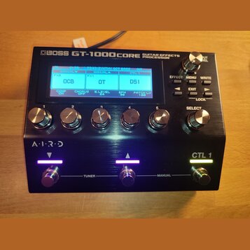 Černý efektový procesor s nápisem BOSS GT-1000CORE GUITAR EFFECTS PROCESSOR. Zařízení má digitální displej s modrým podsvícením, několik kovových oto…
