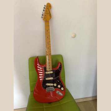 Elektrická kytara typu Stratocaster s červeným bodies, černým pickguardem a světle hnědým hnedem hmatníkem. Na hlavě je nápis "Vintage Modified Strat…