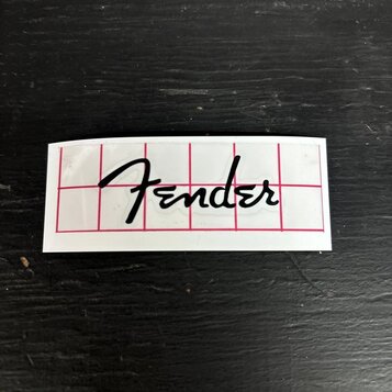 Bílý obdélníkový štítek s černým nápisem Fender v kurzívě. Povrch je proříznut červenou mřížkou s tenkými čarami. Štítek je umístěn na tmavém, textur…