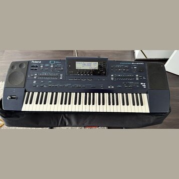 Tmavě modrý digitální klavír značky Roland, model EM-2000. Na horním panelu jsou vidět nápisy Roland, EM-2000 a Creative Keyboard. Nástroj má standar…