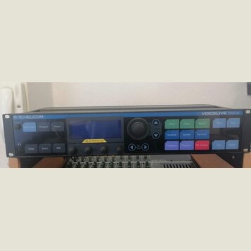 Černý rackový procesor s nápisy TC-HELICON vlevo nahoře a VOICELIVE RACK vpravo nahoře. Zařízení má centrální displej, který je překryt ochrannou fól…