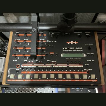 Šedý analogový drum synthesizer značky jomox, model XBASE 999. Zařízení má mnoho ovládacích knoflíků, posuvníků a bílá tlačítka v dolní části s popis…