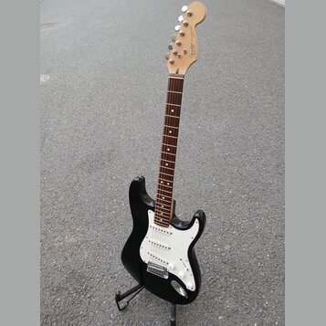 Černá elektrická kytara s bílým pickguardem a hnědým hmatníkem, umístěná na černém stojanu. Na hlavě je nápis Fender STRATOCASTER MADE IN MEXICO. Pov…