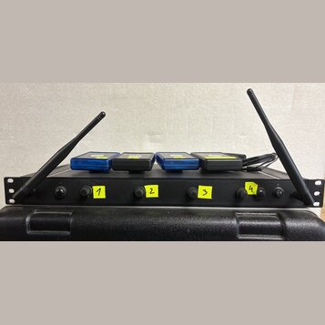 Černý rackový bezdrátový přijímač s dvěma anténami po stranách. Na předním panelu jsou čtyři ovládací prvky označené žlutými samolepkami s čísly 1, 2…