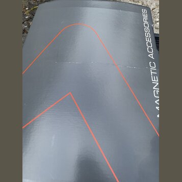 Detailní záběr na šedý povrch s nápisem MAGNETIC ACCESSORIES. Na povrchu jsou vidět tenké červené linky tvořící geometrické tvary. Materiál vykazuje…