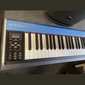 Digitální piano v modré barvě s bílými a černými klávesami. Na horní liště je bílý nápis DEXIBELL a v ovládacím panelu nápisy VIVO S1, Grand Piano Mo…