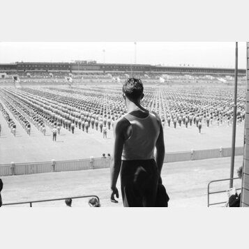 Na fotografii není žádný hudební nástroj ani příslušenství. Snímek zobrazuje muže v tílku zády k fotografu, který se dívá na rozsáhlý shromážděný dav…