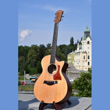 Akustická kytara s cutawayem v přirozené dřevěné barvě, umístěná na černém stojanu. Na hlavě hmatníku je bílý nápis Taylor. Kytara má hnědý hmatník,…