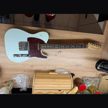Světle modrá elektrická kytara typu Telecaster s červeně hnědým vzorovaným pickguardem a hnedým hmatníkem. Na hlavě kytary je vidět nápis Fender TELE…