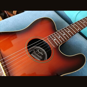 Akustická kytara s barevným přechodem od oranžové k tmavě hnědé (sunburst). Uvnitř ozvučovacího otvoru je štítek s nápisy Fender, Telecoustic, ELECTR…