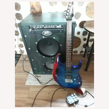 Modrá elektrická kytara s tmavým hnedým hmatníkem a červeným popruhem. Kytara stojí před černým kytarovým kombem, které má v horní části nápis "ROCK…
