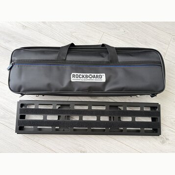 Černý textilní softcase s rukojetmi a modrým detailem u zipu. Na přední straně je bílý štítek s nápisem ROCKBOARD® by WARWICK. Pod pouzdrem leží čern…