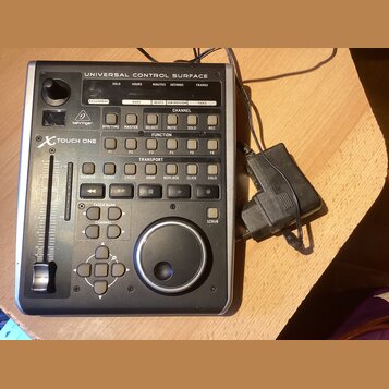 Šedý kontrolní povrch s nápisem UNIVERSAL CONTROL SURFACE a logem behringer. Na levé straně je vidět označení X-TOUCH ONE, jeden posuvník a ovládací…