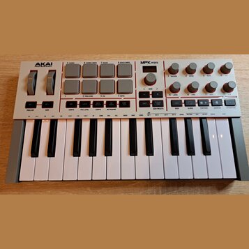 Bílá MIDI klaviatura s názvy AKAI PROFESSIONAL a MPK mini. Horní panel obsahuje šedé čtvercové pady, několik otočných potenciometrů a řadu malých tla…