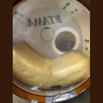 Fotografie zachycuje bílý plastový bubenový kožen s nápisem AMATE a logem značky EVANS, pod kterým je uveden text MADE IN USA. Pod koženem je umístěn…