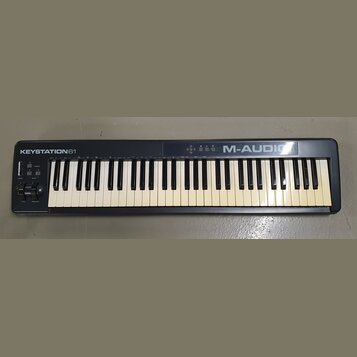 Černá klaviatura s bílými a černými klávesami. Na horní liště je vlevo nápis KEYSTATION61 a vpravo M-AUDIO. V levé části jsou umístěny posuvníky, dva…