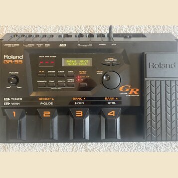 Černý kytarový syntetizátor Roland GR-33 Guitar Synthesizer. Zařízení má digitální displej s nápisem Roland GR-33 Guitar Synth, několik ovládacích tl…
