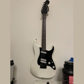 Bílá elektrická kytara s černým hmatníkem a černým pickguardem. Na hlavě je bílý nápis Squier STRATOCASTER. Nástroj má tři snímače, jeden ovládací kn…