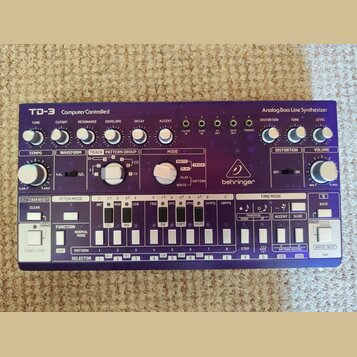 Bílo-fialový analogový syntetizátor s nápisy TD-3 Computer Controlled a Analog Bass Line Synthesizer. Na přístroji je viditelné logo behringer a řada…