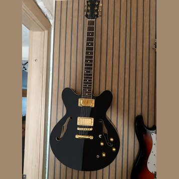 Černá polodubová elektrická kytara s gold hardwarem a hnědým hmatníkem. Nástroj má dva humbuckery, most s laděním strun a tři ovládací knoflíky v zla…