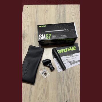 Na dřevěném stole je umístěna krabice a mikrofon značky SHURE s nápisem SM57. Soubor obsahuje černý dynamický mikrofon, černé pouzdro s logem SHURE,…