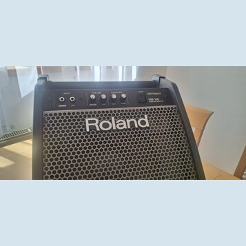 Černý osobní monitor s kovovou mřížkou, na které je bílý nápis Roland. Horní ovládací panel obsahuje vstupy INPUT (V-Drums, LINE), ovladače VOLUME (V…
