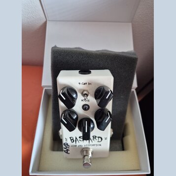 Bílý efekt pedál s nápisy BAS TARD a LOW END DISTORTION v bílé krabičce s šedou pěnovou vložkou. Pedál má čtyři černé oválné ovládací knoflíky, jeden…