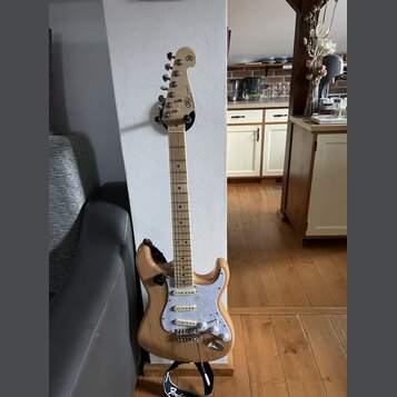Elektrická kytara s přírodním dřevěným povrchem těla a světlou hmatníkovou deskou. Na hlavě je viditelné logo "SG". Kytara má bílý pickguard s textur…