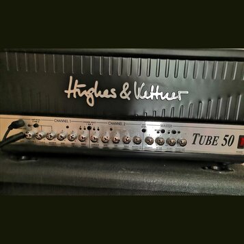 Detail kytarového komba s černým povrchem a stříbrným ovládacím panelem. Na horní části je nápis Hughes & Kettner a vpravo na panelu označení Tube 50…