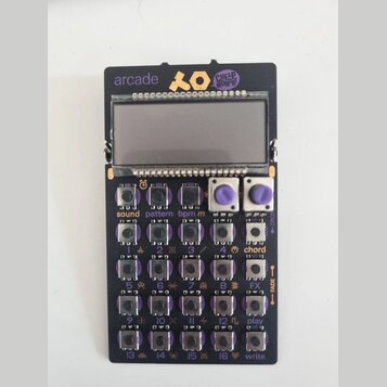 Černý elektronický sekvencer s displejem a ovládacími prvky. V horní části je nápis arcade, logo XO a nálepka CHEAP MONDAY. Zařízení obsahuje řadu tl…